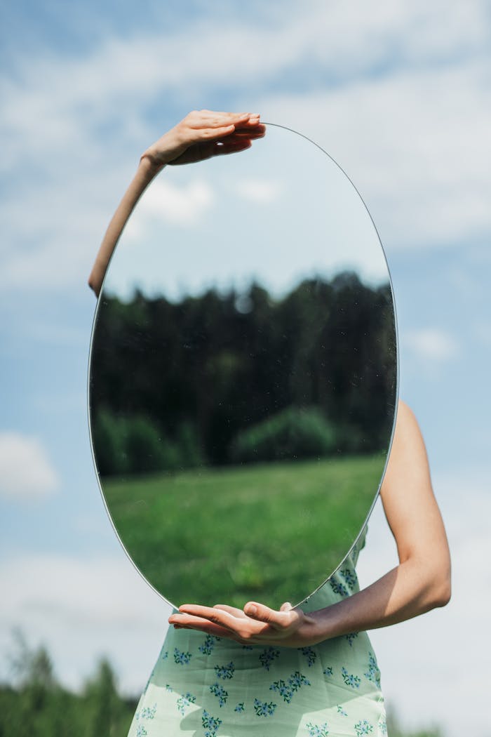 Proč se mi v životě opakují stejné vztahové vzorce? A jak z nich vystoupit A woman in a floral dress holds an oval mirror reflecting a forest on a sunny day.